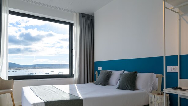 A hotel room with a bed, a chair, a nightstand, and a view of the sea through a large window. - Alda Corrubedo (Ribeira)