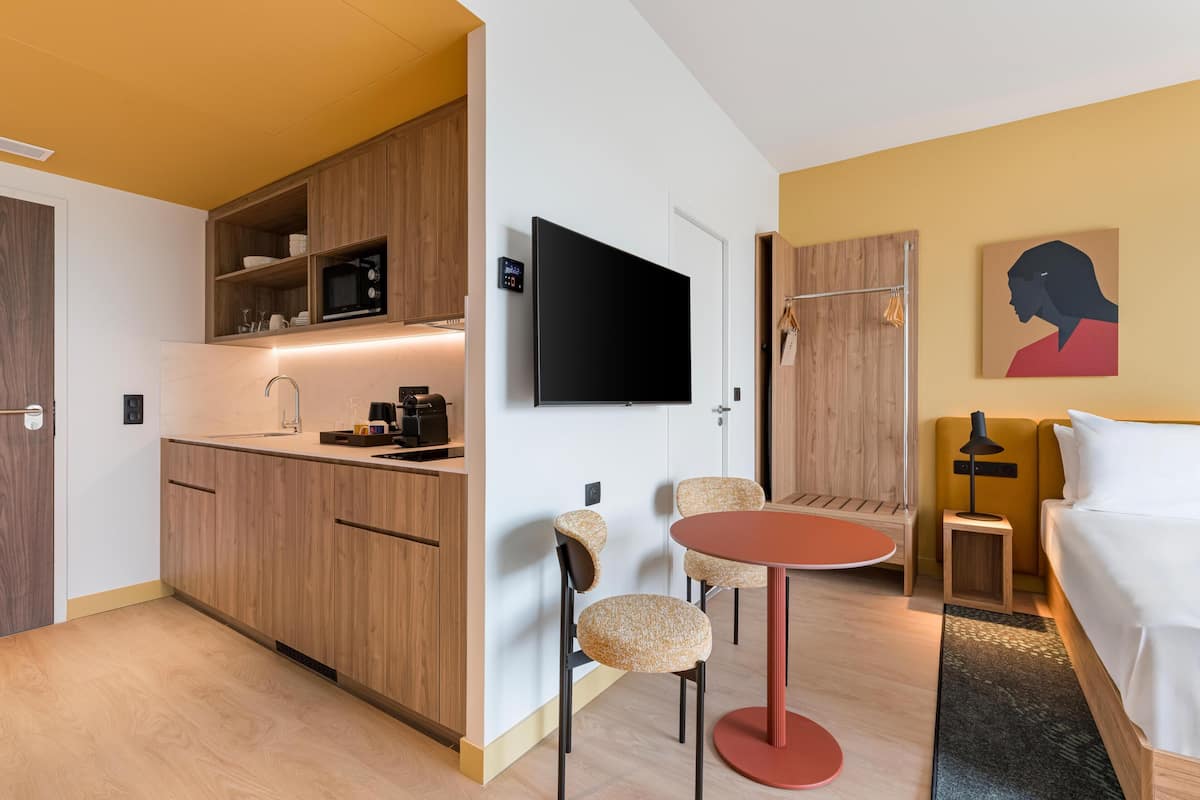 Een kleine hotelkamer met kitchenette, flatscreen-tv, bed, een tafeltje en stoel.