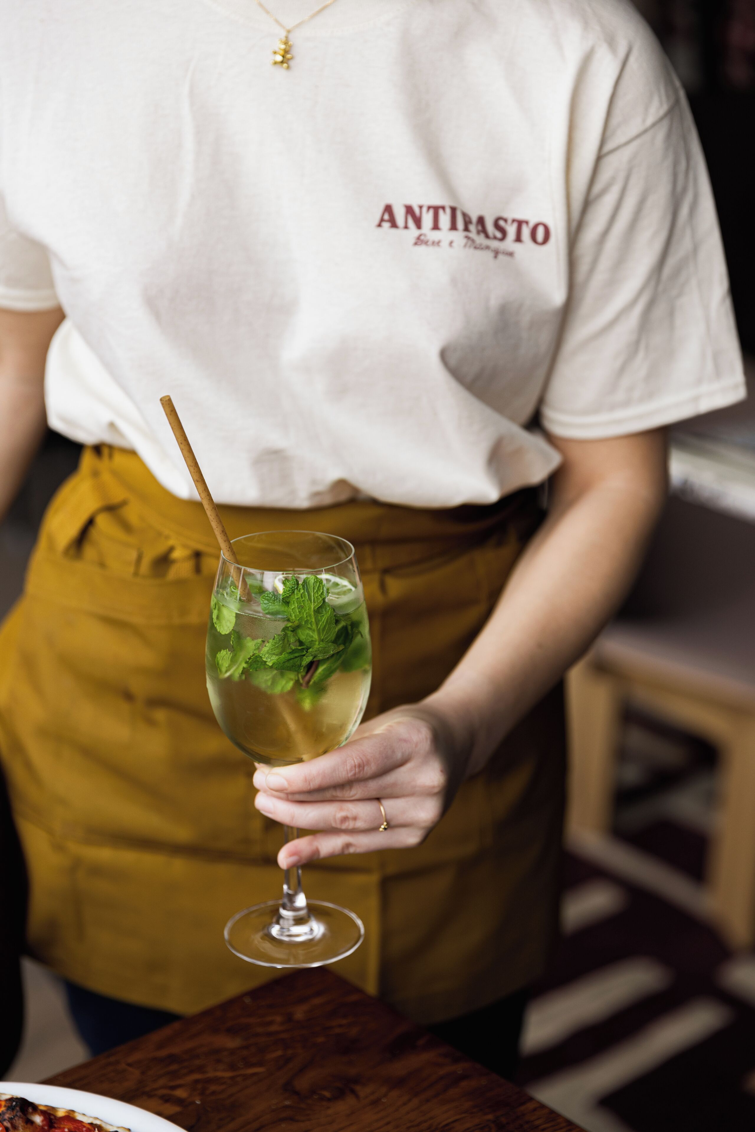 A person wearing a white shirt with "ANTIMASTO" printed on it, holding a glass with a straw and mint leaves.