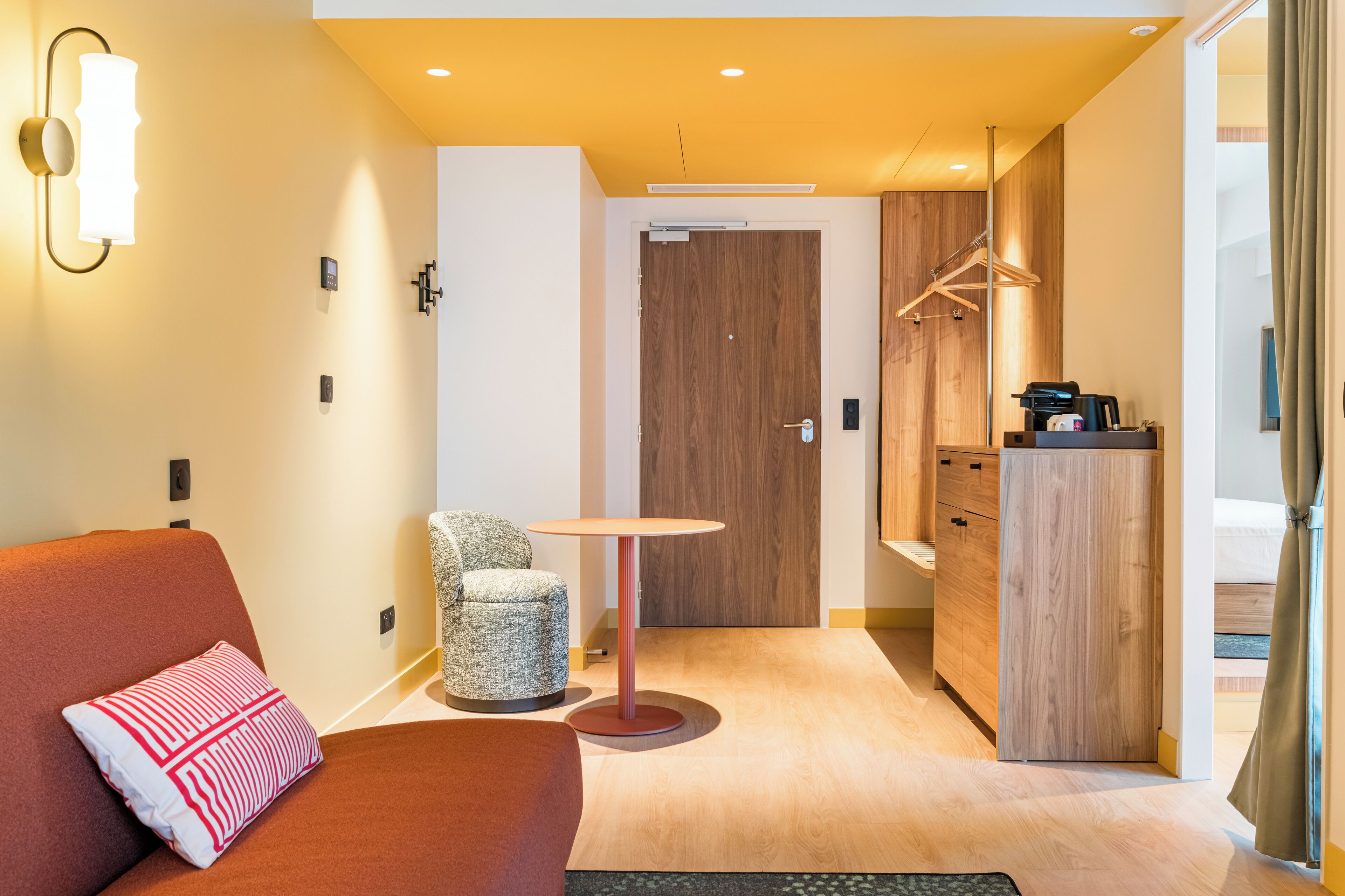 Ein modernes Hotelzimmer mit einer Couch, einem kleinen runden Tisch, einem Stuhl, einem Holzschrank und einer Tür zu einem Badezimmer.
