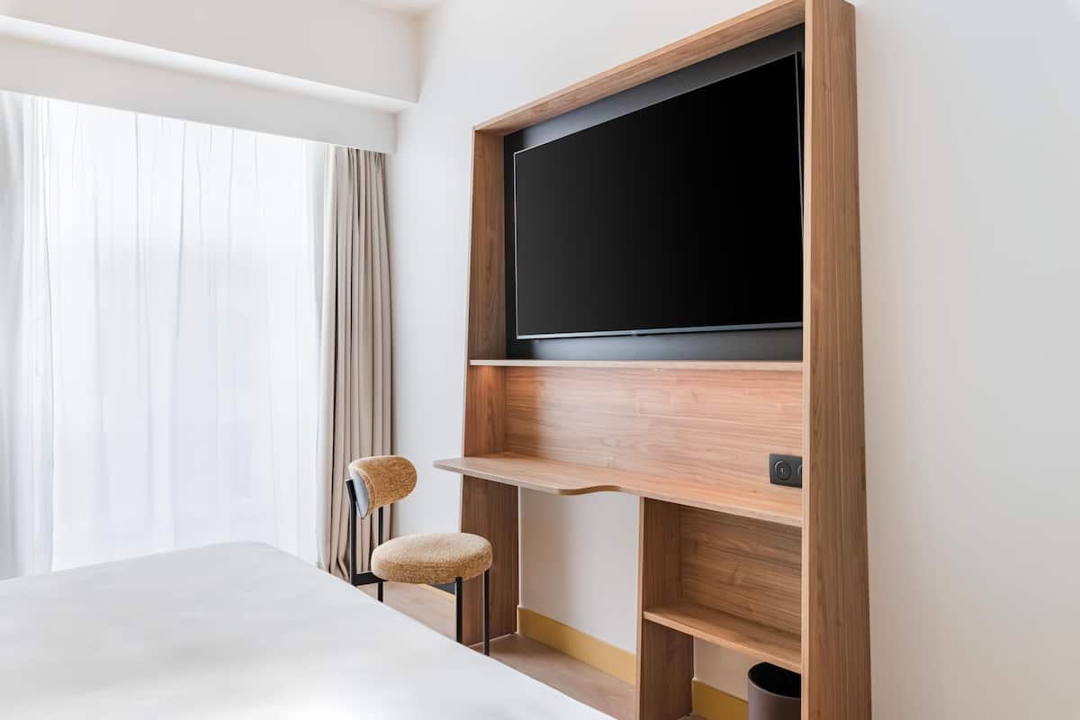 Een hotelkamer met een bed, een televisie bevestigd aan een houten plank, een stoel en een raam met gordijnen.