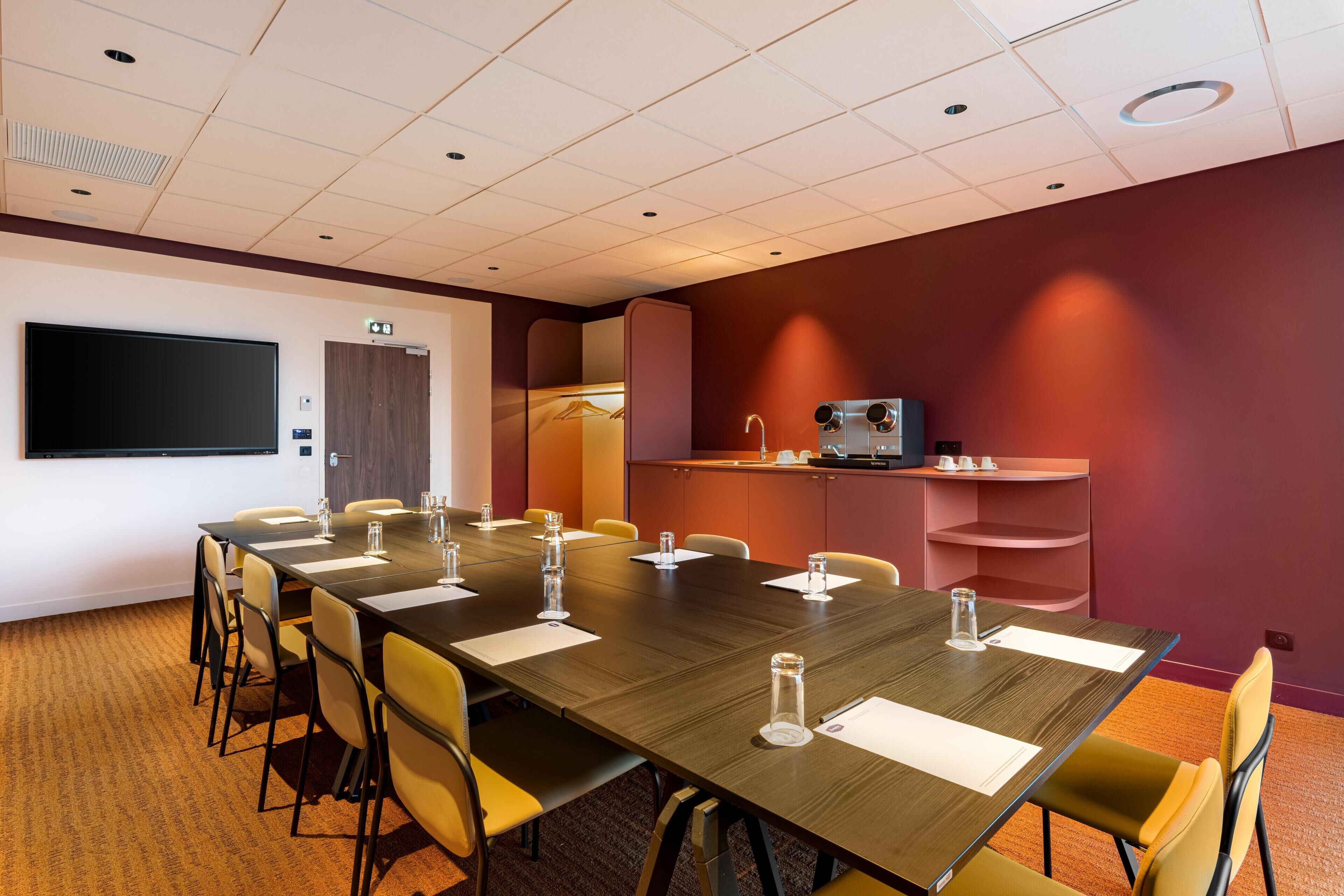 A conference room with a long table, chairs, a projector screen, a coffee machine, and a small kitchenette.