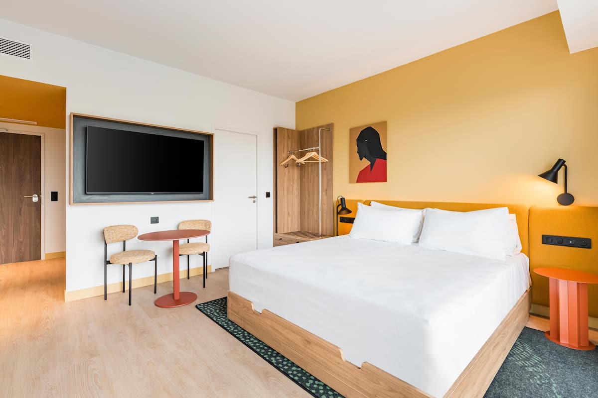 Een moderne hotelkamer met een groot bed, een aan de muur bevestigde televisie, een tafeltje met stoelen, een kledingkast en een schilderij.