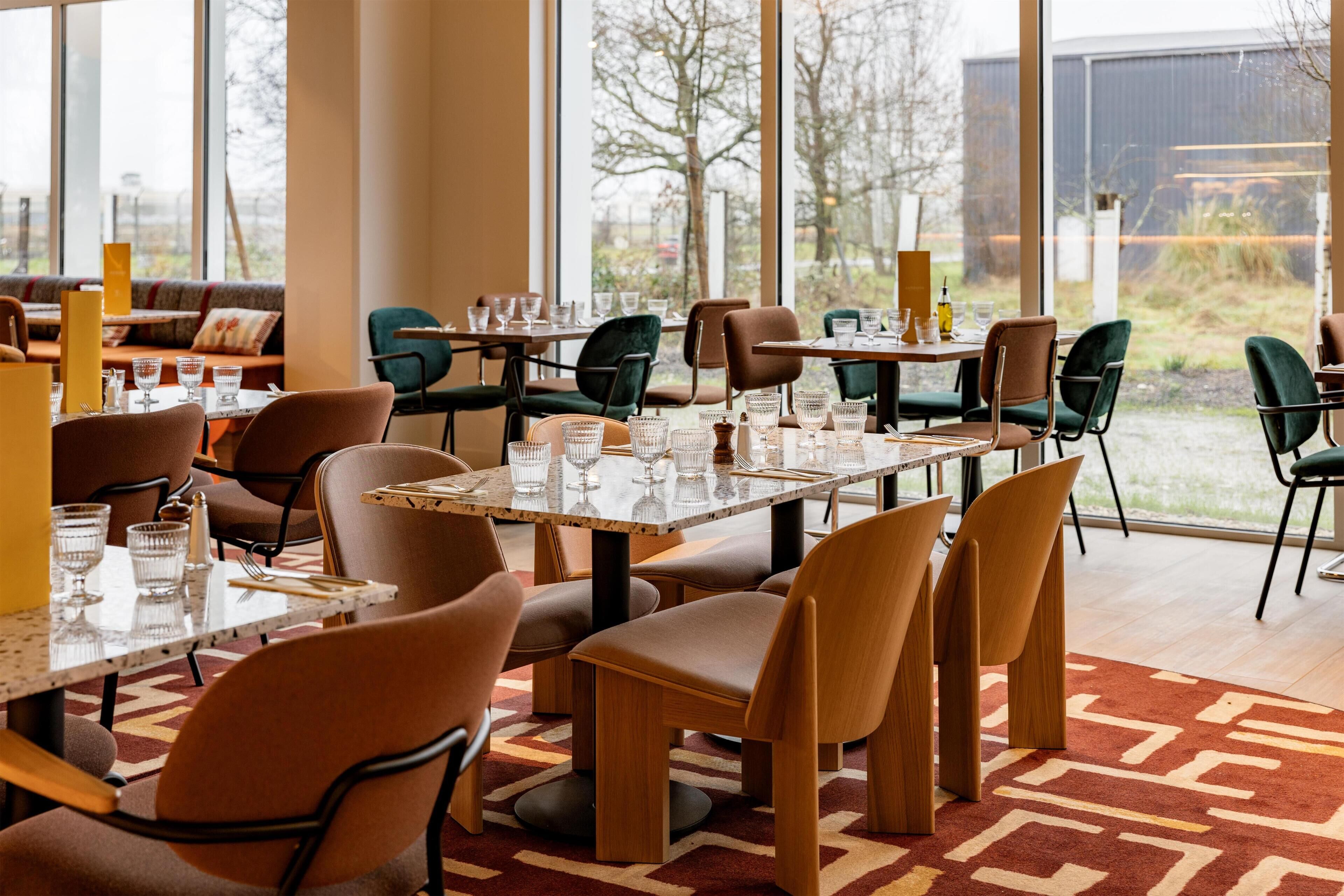 A restaurant with tables and chairs arranged for dining, large windows offering a view of the outdoors, and a patterned carpet on the floor.