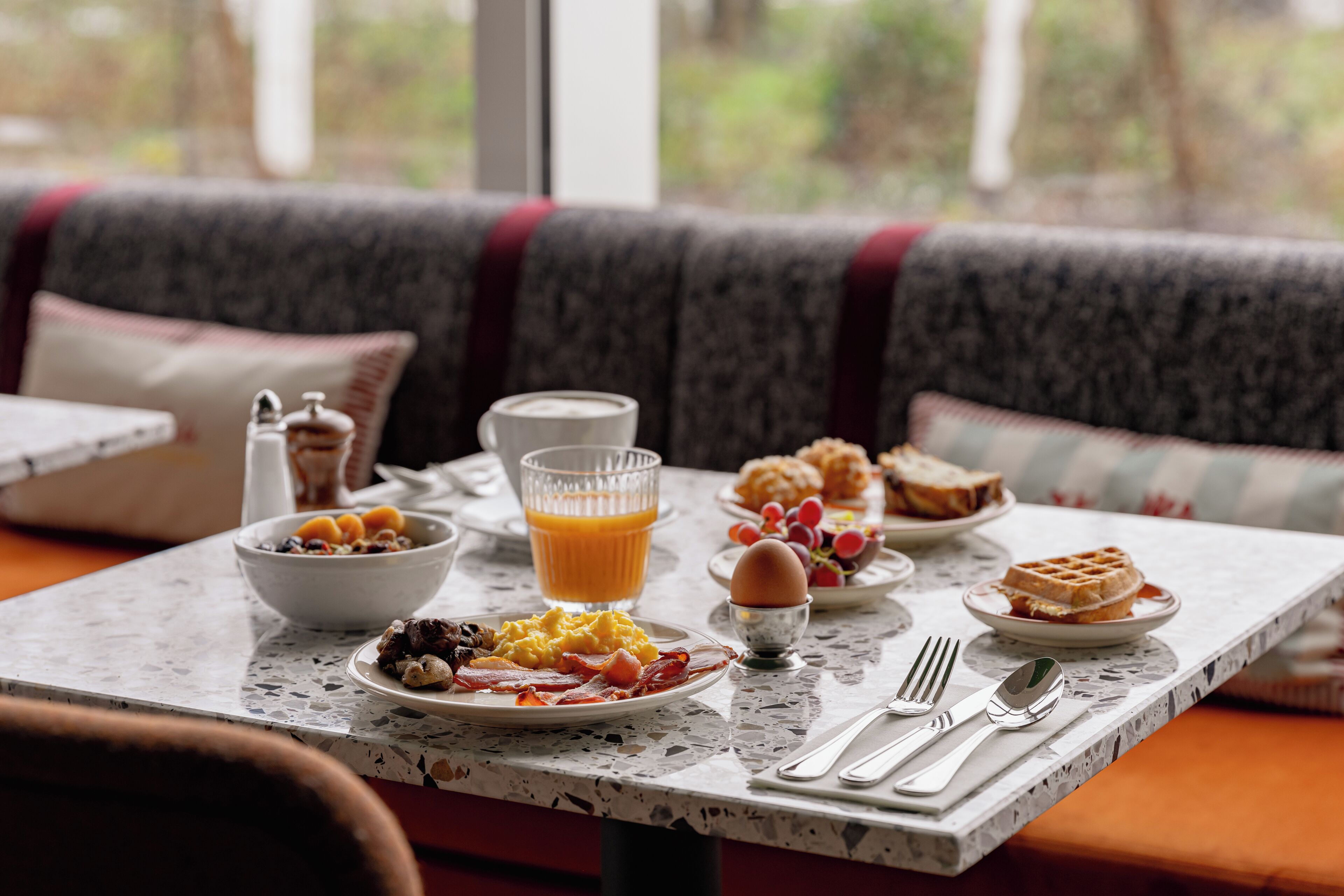 Una mesa de desayuno con platos de huevos, tocino, tostadas y gofres, un vaso de zumo de naranja y un tazón de cereal.