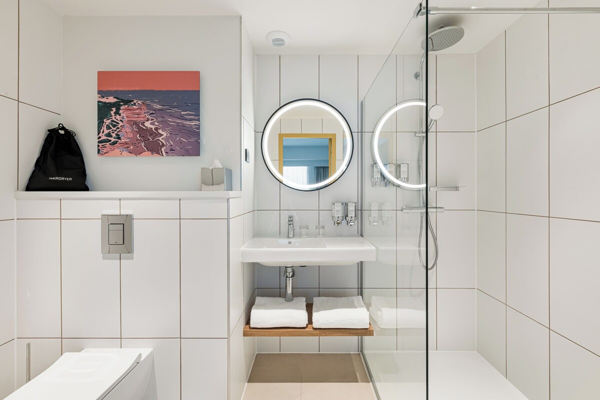 Ein modernes Badezimmer mit einem weißen Waschbecken, zwei Spiegeln, einer Duschkabine aus Glas und einem Handtuchhalter.