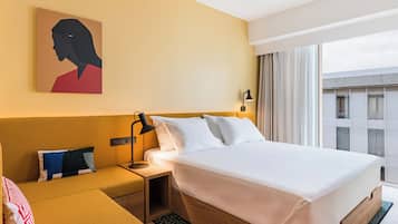 A hotel room with a bed, a nightstand, a lamp, a painting, and a view of the city.