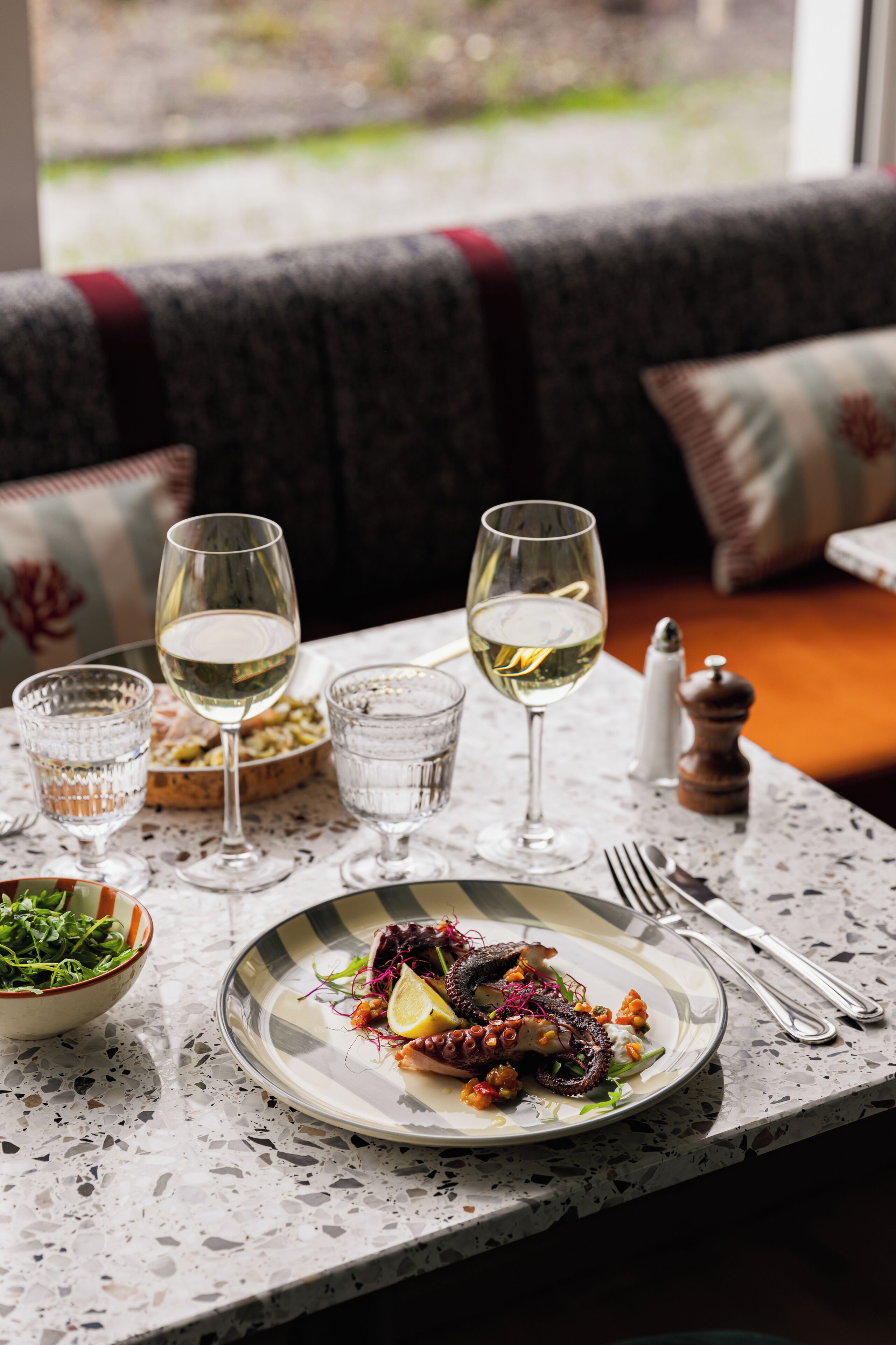 A dining table with a plate of food, glasses of white wine, and a salt and pepper shaker.