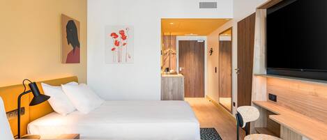 A modern hotel room with a bed, a desk, a TV, and artwork on the walls.