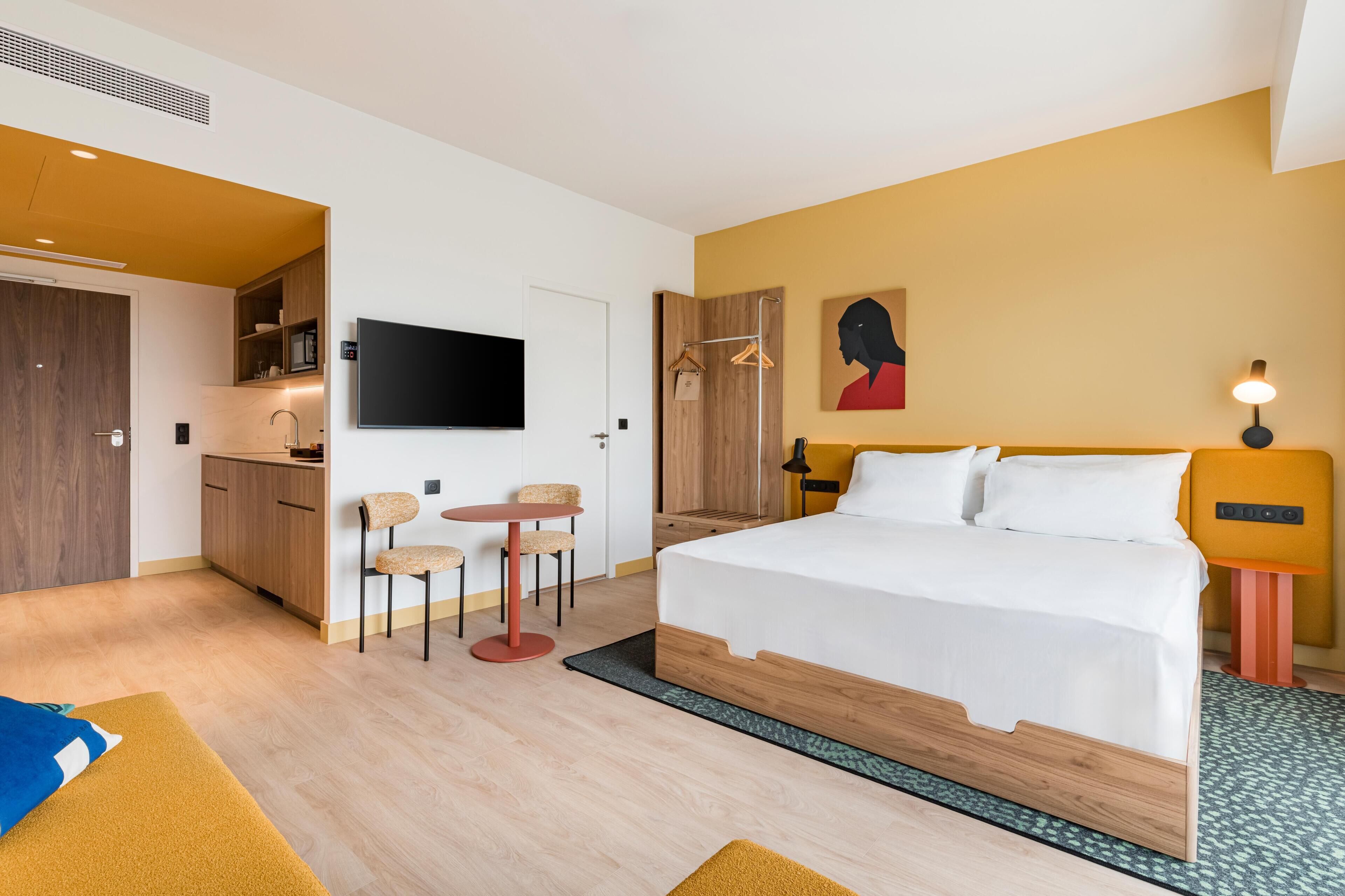 A modern hotel room with a bed, a small kitchenette, a TV, and a painting on the wall.