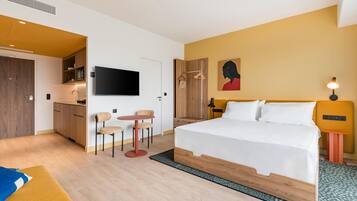 A modern hotel room with a bed, a small kitchenette, a TV, and a painting on the wall.
