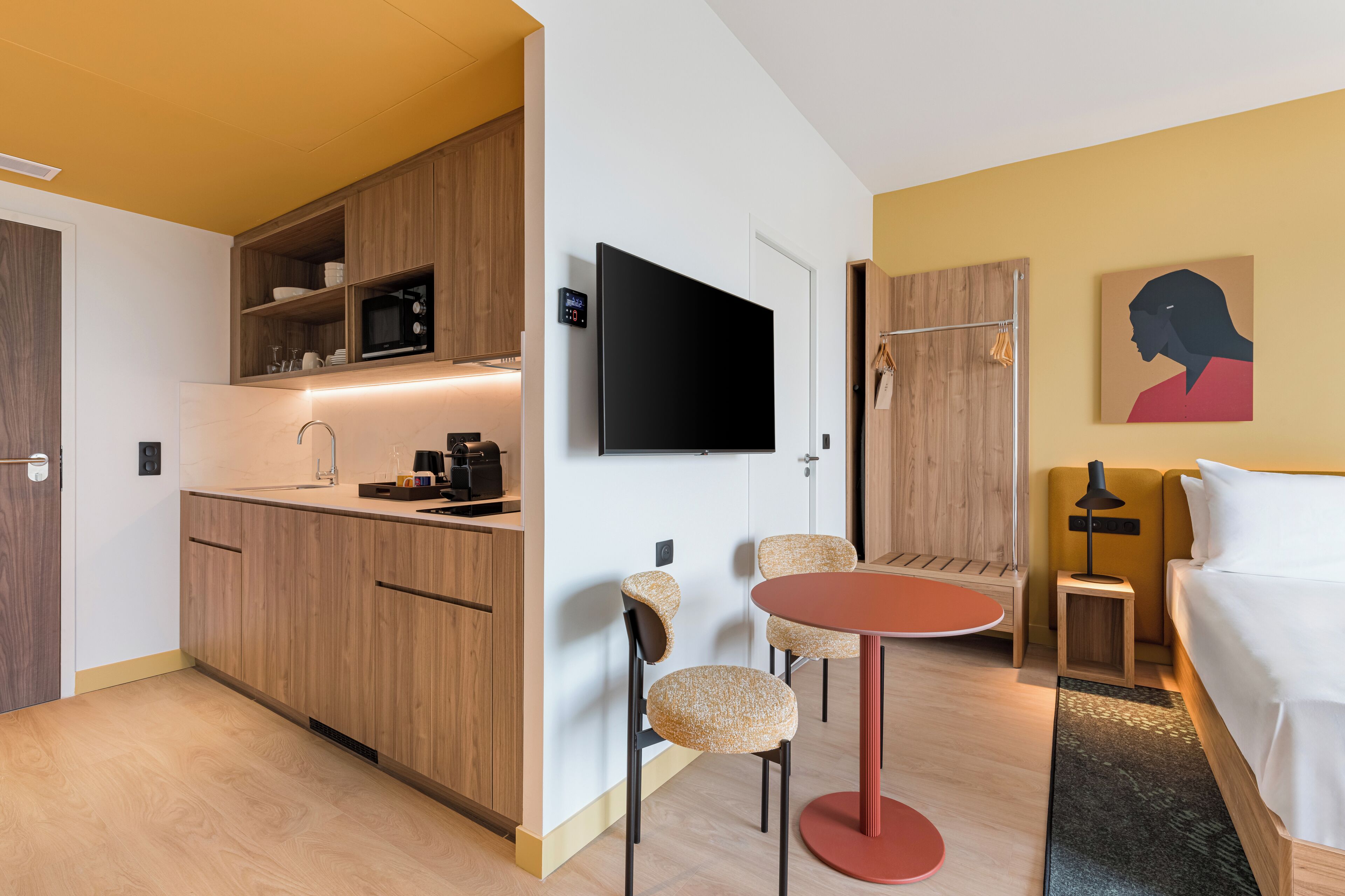 A compact hotel room with a kitchenette, a flat-screen TV, a bed, a small table, and two chairs.