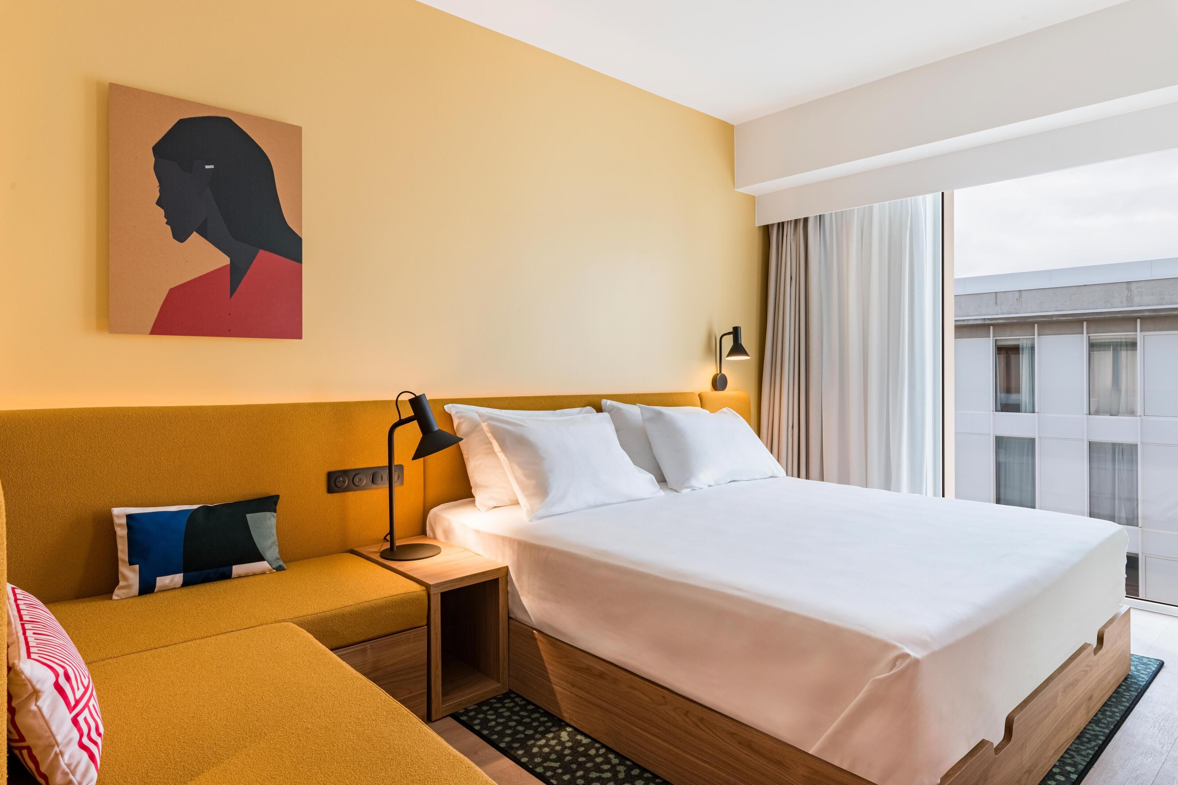 Une chambre d’hôtel avec un lit, une table de chevet, une lampe, un tableau et une vue sur l’extérieur.
