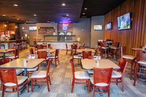 A restaurant with multiple tables and chairs, a counter with a menu board, and a television screen displaying a sports event. - Hope Hotel & Conference Center (Dayton)