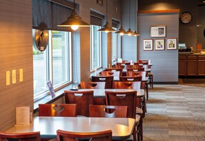 A restaurant with wooden tables and chairs, pendant lighting, large windows, and framed pictures on the wall.