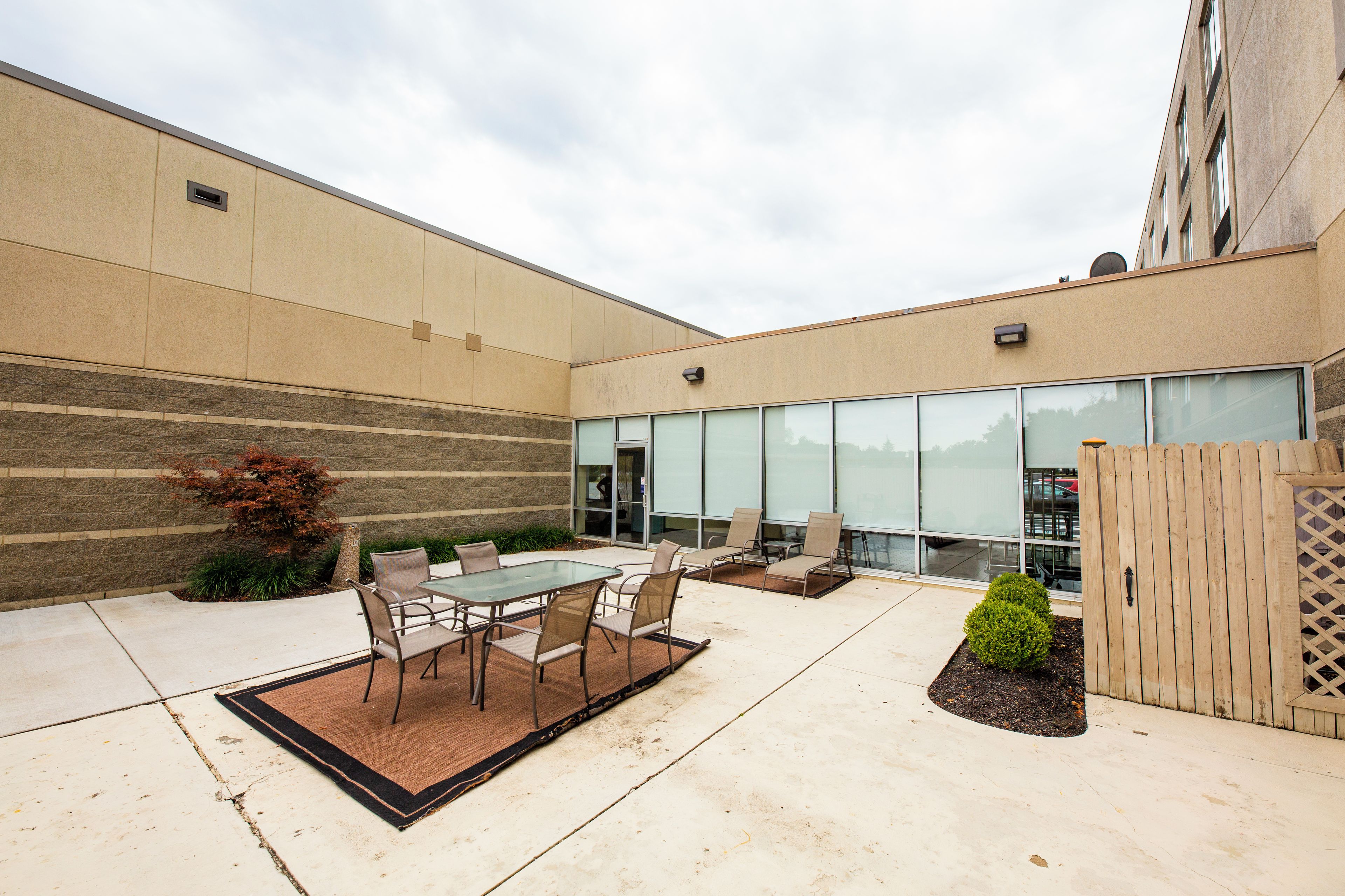 An outdoor patio area with a glass table and chairs, a small garden, and a building with large windows in the background.