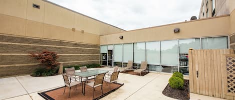 An outdoor patio area with a glass table and chairs, a small garden, and a building with large windows in the background.