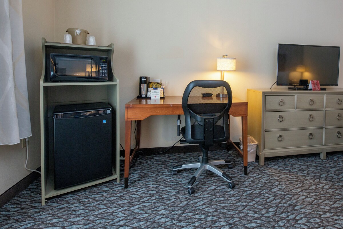 A hotel room with a desk, a microwave, a refrigerator, a chair, a TV, a lamp, and a dresser.