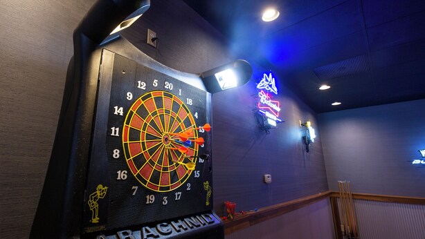 A dartboard with three darts, a neon sign, and a wall-mounted light fixture. - Hope Hotel & Conference Center (Dayton)