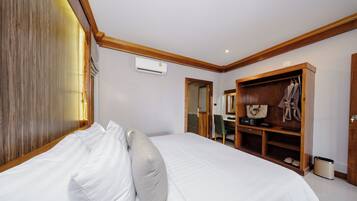 A bedroom with a large bed, a wooden wardrobe, a desk with a chair, and an air conditioning unit.