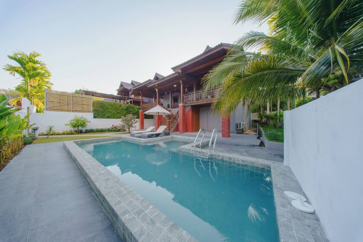 Una piscina con bordo piastrellato, un'area relax coperta con ombrelloni e un edificio con tetto rosso e colonne in legno.