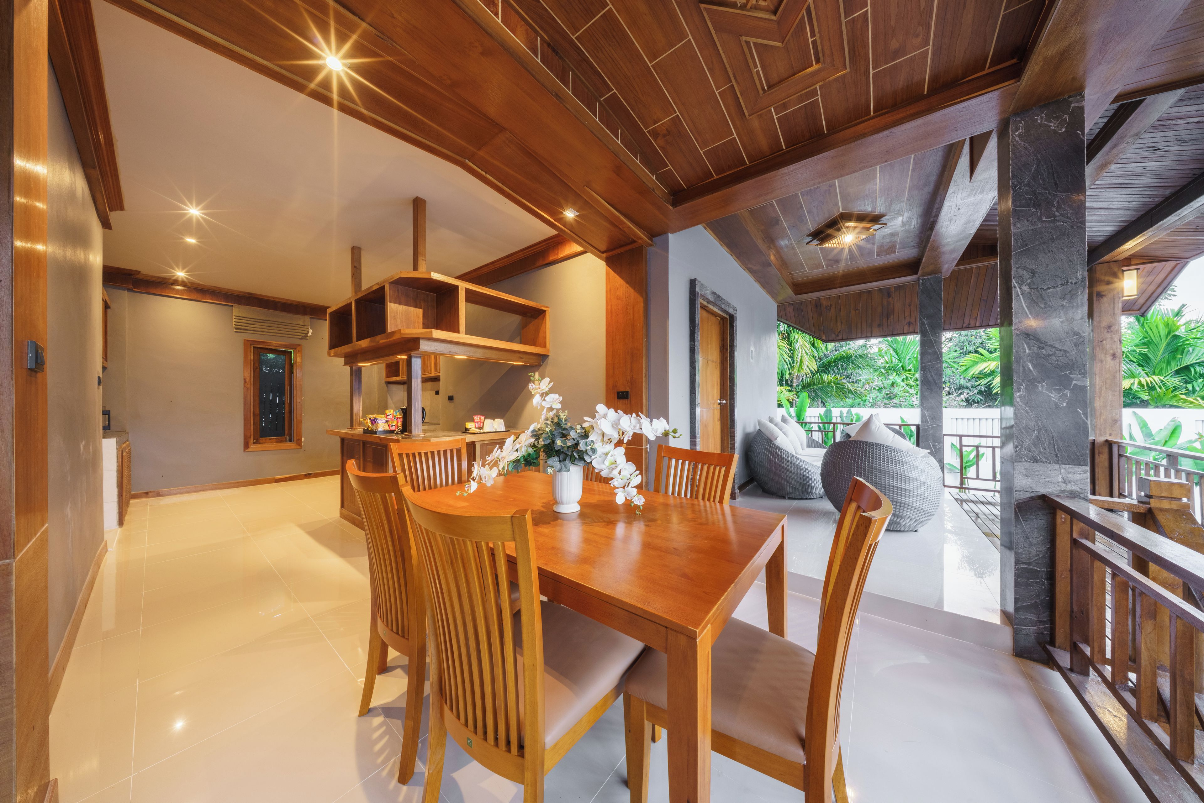 A spacious dining area with a wooden table and chairs, a white vase with flowers, and a view of lush greenery outside.