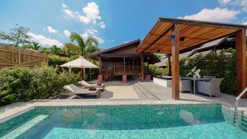 A swimming pool with a wooden deck and lounge chairs, surrounded by lush greenery and a clear blue sky.