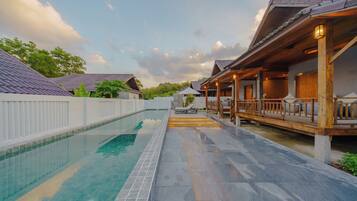 A swimming pool with a tiled deck, wooden deck with railings, and a covered area with seating.