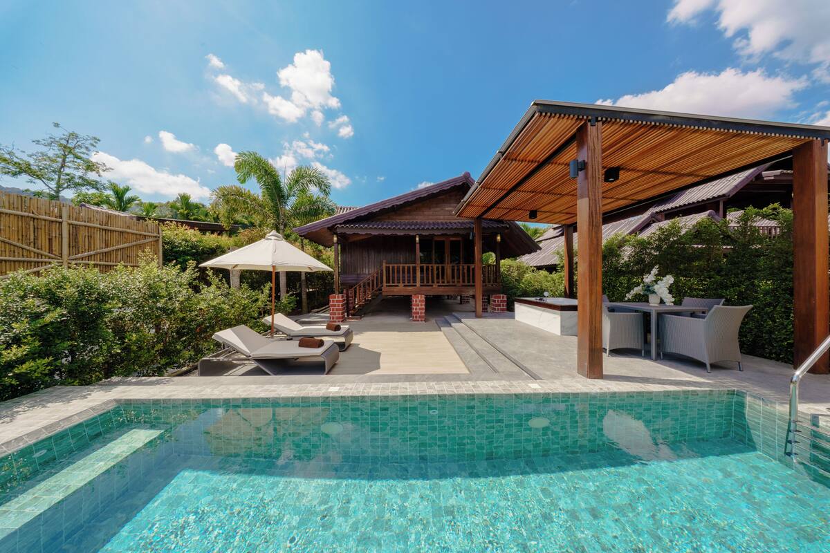 Una piscina con bordo in legno e lettini, circondata da vegetazione rigogliosa e un cielo azzurro.
