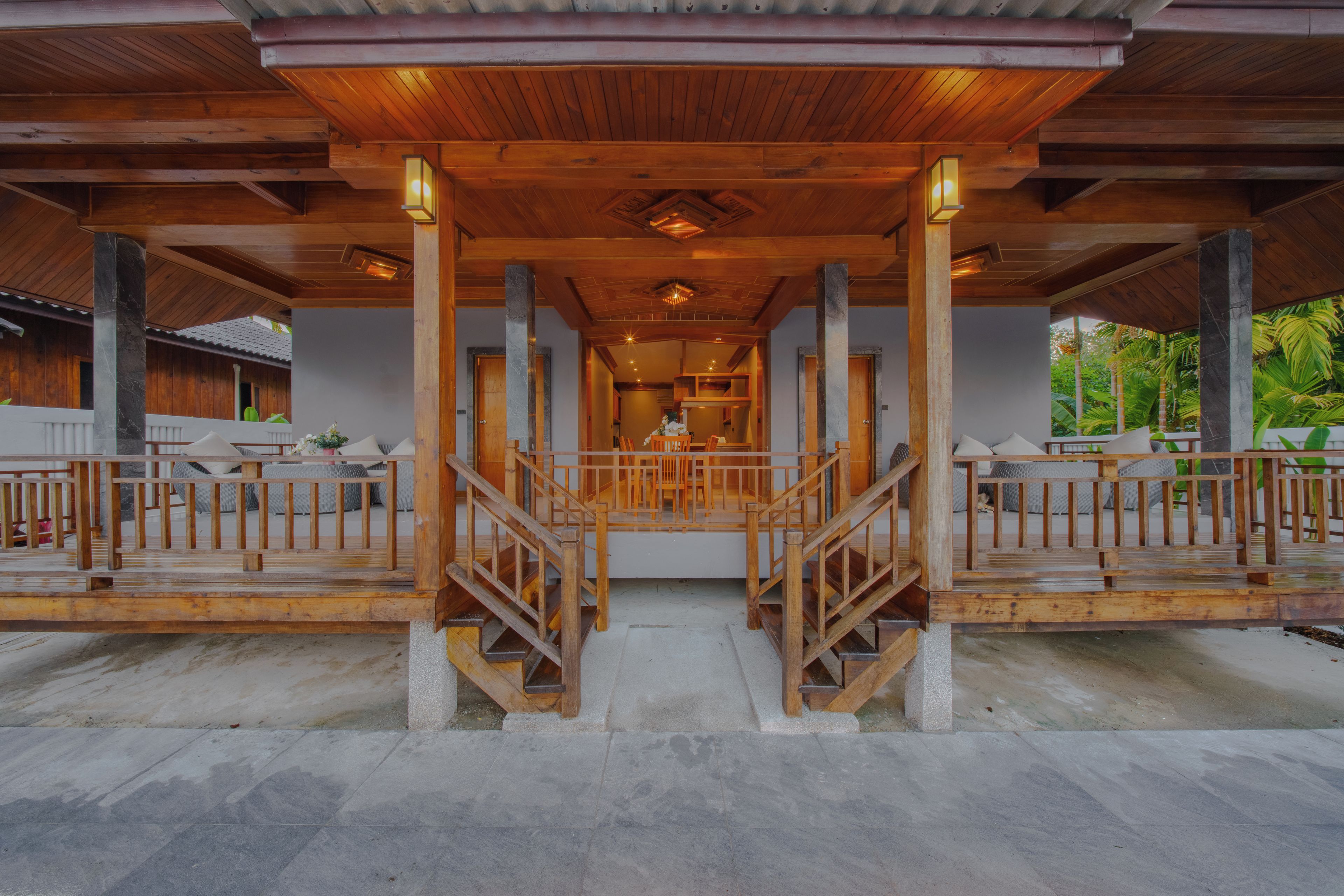 A wooden covered walkway with a central staircase leading to a covered area, surrounded by a white fence and greenery.