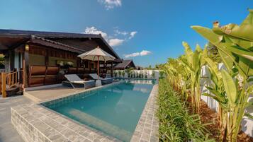 A swimming pool with lounge chairs, a wooden structure with a tiled roof, and lush greenery.