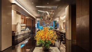 Un moderno vestíbulo de hotel con un arreglo floral central, una recepción y un área de comedor al fondo.