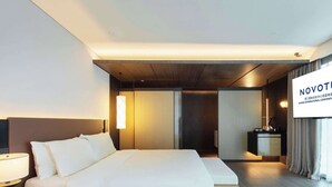 Una habitación de hotel moderna con una cama grande, una mesita de noche y una pantalla de televisión que muestra 'NOVOTEL'.