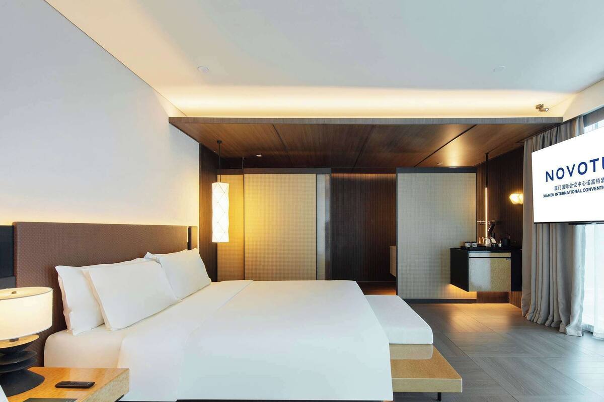 Ein modernes Hotelzimmer mit einem großen Bett, einem Nachttisch und einem Fernseher, der 'NOVOTEL' anzeigt.