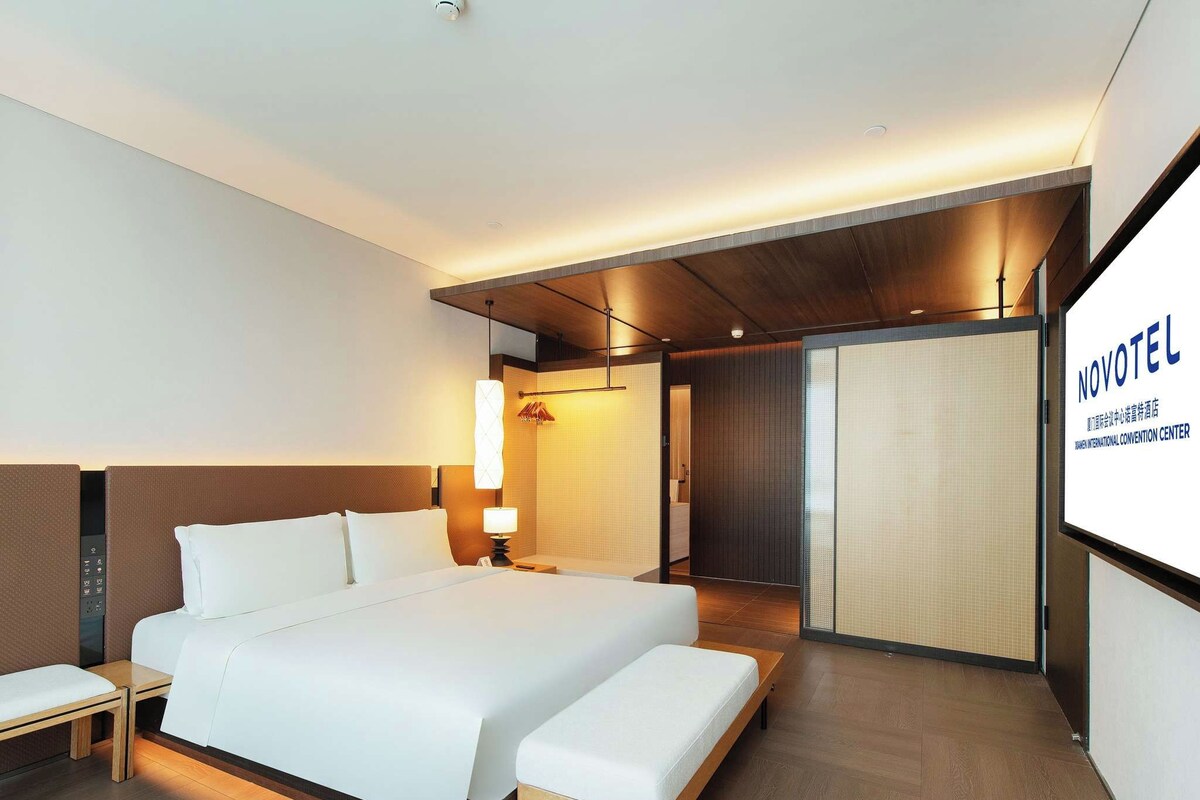 A modern hotel room with a large bed, bedside tables, a desk, and a view of the Novotel International Convention Center.