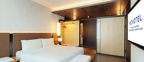 A modern hotel room with a large bed, bedside tables, a desk, and a view of the Novotel International Convention Center.