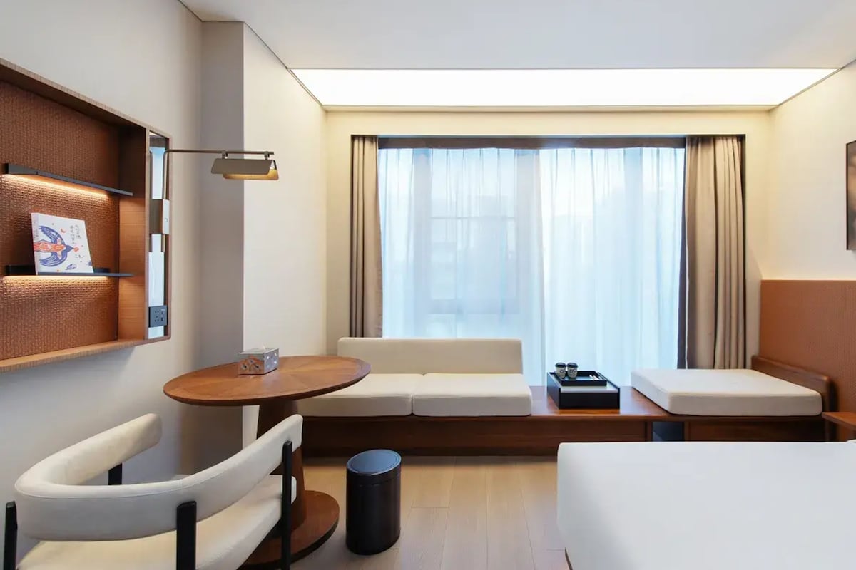 Ein modernes Hotelzimmer mit einer Couch, einem kleinen Tisch, einem Sessel, einem Nachttisch mit Lampe und einem Vorhangfenster.