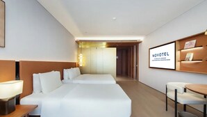 Habitación de hotel con una cama grande, mesitas de noche, un escritorio y una silla. Se ve un cartel en la pared y un mostrador de recepción al fondo.