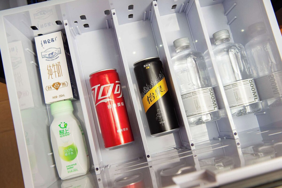 Ein Kühlschrank mit verschiedenen Getränken und Behältern, darunter eine Kokosnusswasserflasche, eine rote Dose und mehrere durchsichtige Flaschen.