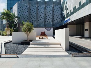 A modern hotel entrance with a geometric facade, a bench, and a small tree.