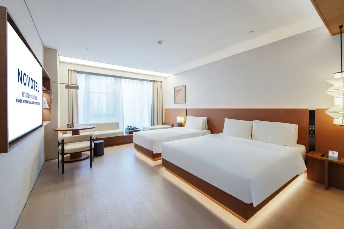 Ein Hotelzimmer mit zwei Betten, einem Schreibtisch, einem Stuhl, einem kleinen Tisch, einer Lampe und einem Fenster mit Vorhängen.