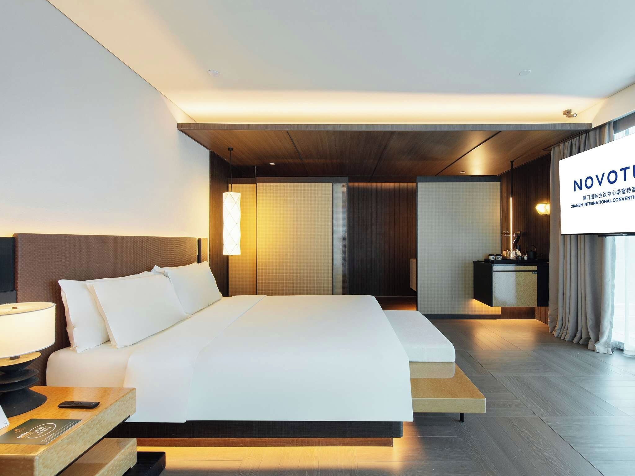A modern hotel room with a large bed, bedside table, and a TV screen displaying 'NOVOTEL'.