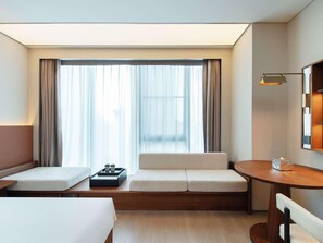 A hotel room with a sofa, a small table, a bedside table, and a large window with curtains. - Novotel Xiamen International Convention Center (Xiamen)