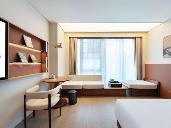 A hotel room with a bed, a sofa, a chair, a small table, a trash bin, and a shelf with books. - Novotel Xiamen International Convention Center (Xiamen)