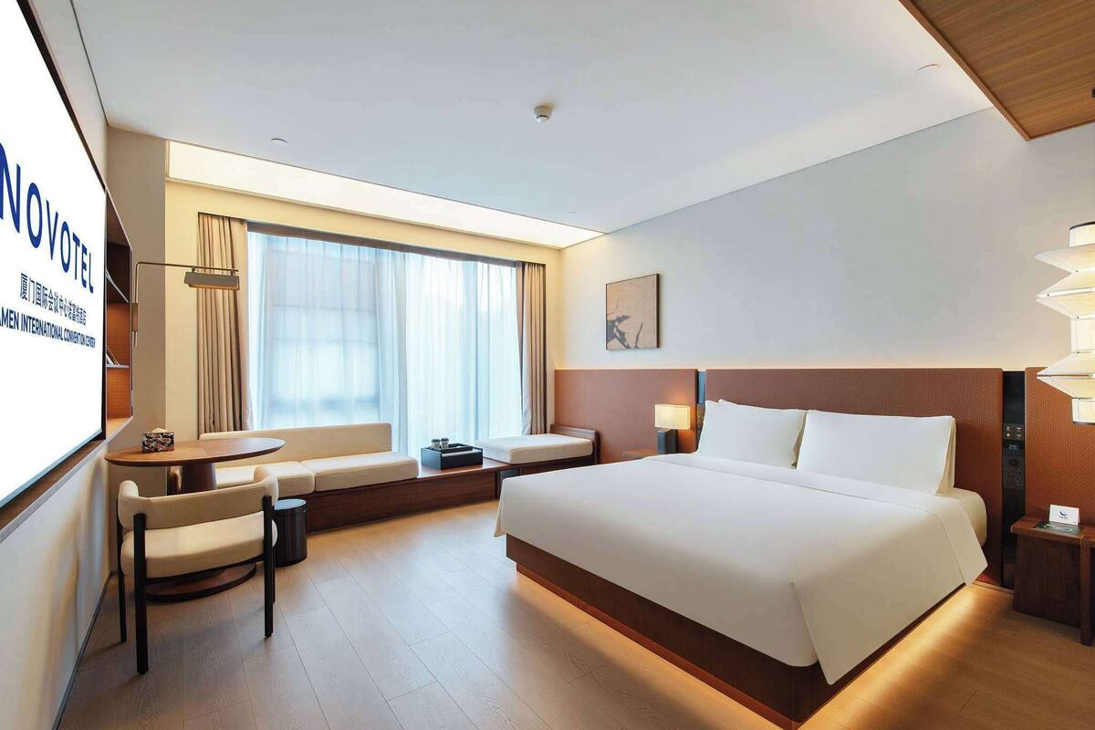 Ein Hotelzimmer mit einem großen Bett, einer Sitzecke mit Stühlen, einem kleinen Tisch und einem großen Fenster mit Vorhängen.