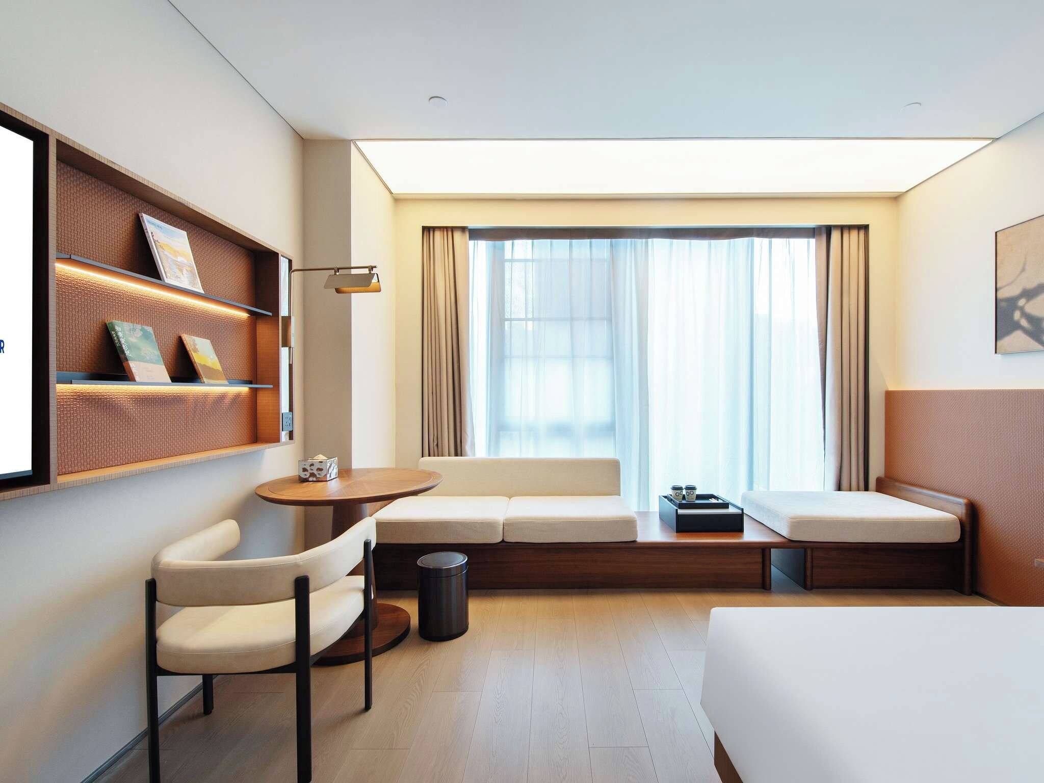 A hotel room with a sofa, a chair, a small table, a trash bin, and a bed.