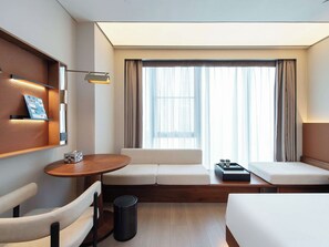 Ein modernes Hotelzimmer mit Schlafsofa, kleinem Holztisch, Stuhl, Mülleimer und einem Fenster mit Vorhängen.