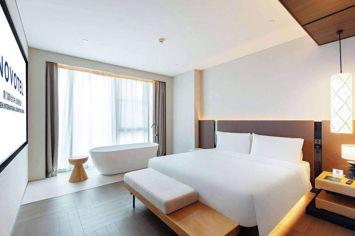 Ein modernes Hotelzimmer mit einem großen Bett, einer eingebauten Sitzbank, einer Badewanne und einem Nachttisch mit Lampe.