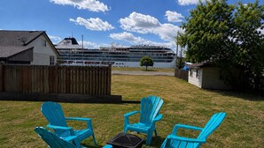 A grassy yard with four blue Adirondack chairs, a black suitcase, and a wooden fence. In the background, a cruise ship named 'Viking' is docked near a body of water.