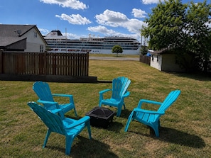 A grassy yard with four blue Adirondack chairs, a black suitcase, and a wooden fence. In the background, a cruise ship named 'Viking' is docked near a body of water.